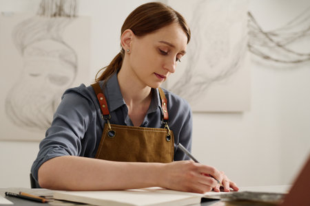 Young pensive craftswoman with pencil in hand looking at page of notepad while thinking of new creative ideasの写真素材