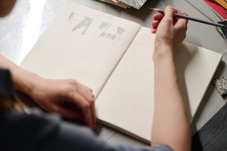 High angle of hands of young unrecognizable craftswoman with pencil over sketches on page of open...
