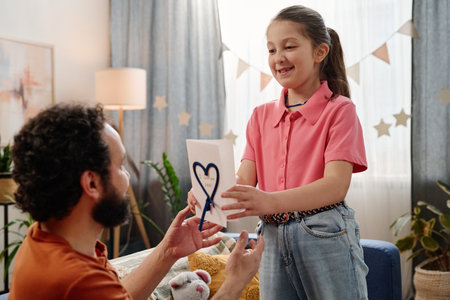 Man gifting card to smiling girl in cozy living room decorated with stars and plants, child wearing pink shirt and jeans, teddy bear on couch, light streaming through curtainsの写真素材