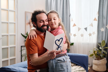 Father and daughter sharing emotional moment while holding greeting card together in cozy living...