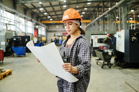 Engineer in hard hat examining blueprint inside spacious industrial manufacturing facility with modern equipment aroundの写真素材