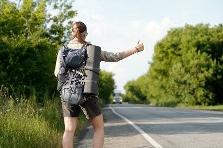 Backpack and rolled sleeping mat on back of young female hitchhiker standing by highway and catching vehicleの写真素材