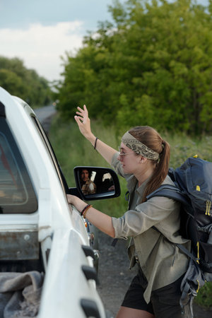 Young pretty woman with backpack standing by white pickup truck and asking driver if he takes her to...