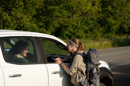 Young female hitchhiker with backpack standing by car window and talking to vehicle driver while...