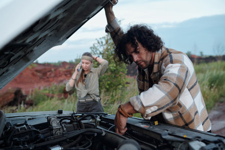 Young man with dark wavy hair bending over open hood of pickup truck and checking engine against his girlfriend calling repair serviceの写真素材