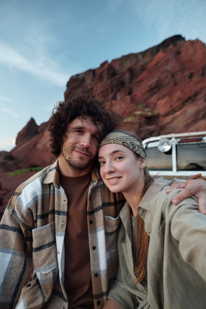 Happy young female tourist stretching her hand with smartphone and taking selfie with her affectionate boyfriend during tripの写真素材