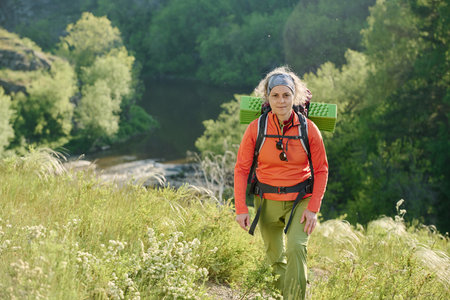 Active mature female camper with backpack looking straight while walking down forest path among green grass during hiking tripの写真素材