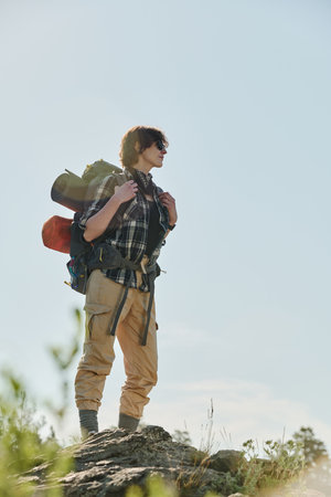 Full length portrait of young active woman with backpack standing on mountain top against bright blue sky and enjoying summer adventureの写真素材