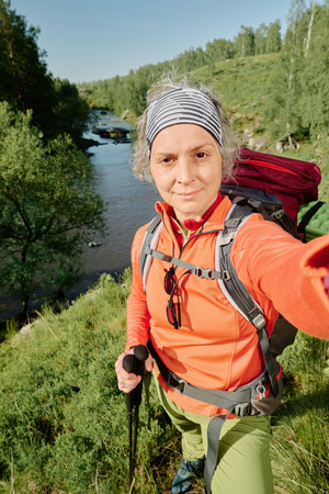 Mature female backpacker in activewear looking at camera while taking selfie against riverside...