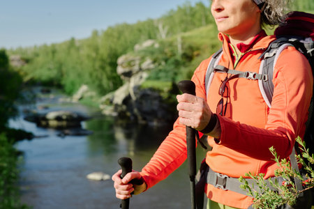 Cropped shot of active female hiker with trekking sticks walking in front of camera while passing by natural landscape with riversideの写真素材