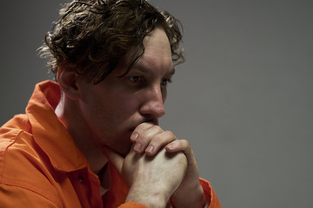 Contemplative man wearing orange jumpsuit resting chin on hands, pensive expression seen on his face...