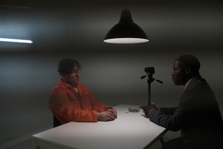 Man wearing orange jumpsuit sitting at table during interrogation session facing another man in suit both men deep in conversation under bright lightの写真素材