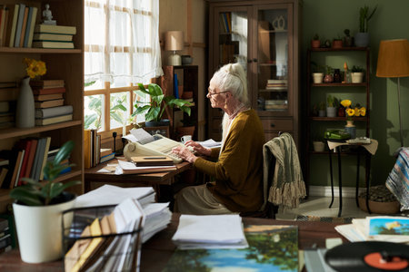 Elderly woman sitting by window reading documents in cozy room with lots of books and plants around, creating a serene atmosphere for thoughtful reflectionの写真素材