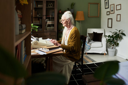 Elderly woman wearing glasses sitting at desk, engrossed in reading a book, surrounded by books and plants in well-decorated home study area with warm ambianceの写真素材