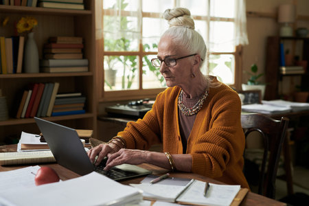 Elderly woman with white hair in a bun working on laptop in a home office. Books, notebooks, and...