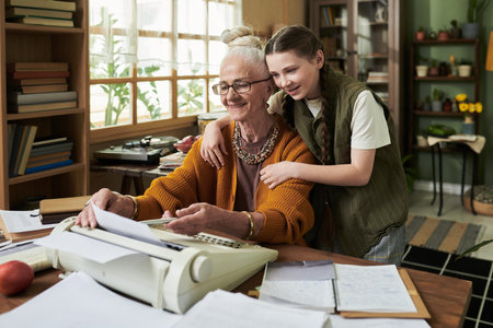 Elderly woman typing on typewriter while young girl hugging her from behind in cozy home office setting. Bonding moment between senior and child during relaxed afternoonの写真素材