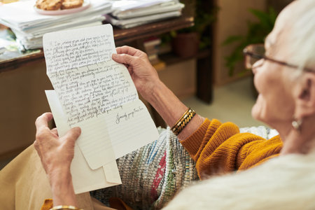 Elderly person reading an intricately handwritten letter, sitting beside a cozy desk filled with stacked papers and plants, expressing emotions of nostalgia and longing while reminiscingの写真素材