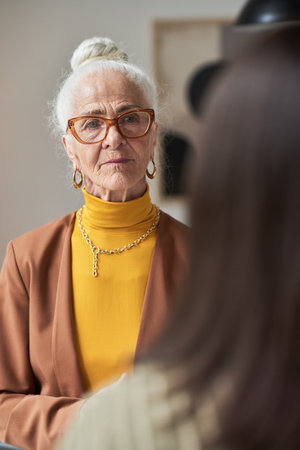 Portrait of elderly woman with white hair in bun, wearing glasses and golden jewelry, standing indoors, looking straight at camera with a serious expressionの写真素材