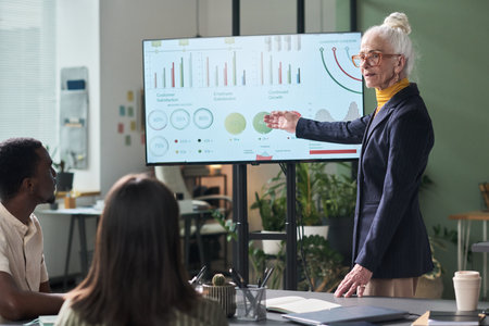 Senior businesswoman presenting data analysis on large screen to diverse team in modern office...