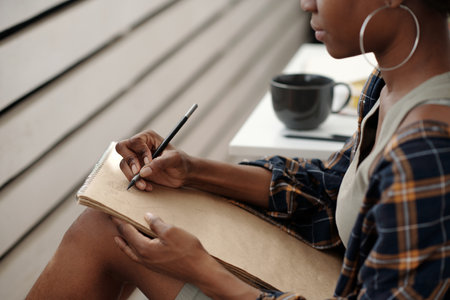Person holding pencil writing in notebook while sitting at outdoor setting with coffee mug on table nearby. Wearing casual outfit featuring large hoop earringsの写真素材