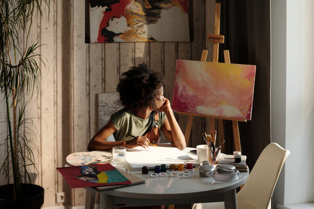Painter working on canvas in bright studio with various art supplies and paintings around. Light streaming through window, adding illumination to creative spaceの写真素材