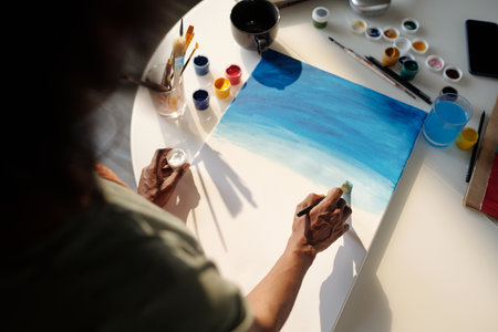 Person painting blue hues on canvas among art supplies including brushes, paint containers, and a glass of water Creating abstract art with soft strokes and blending colorsの写真素材
