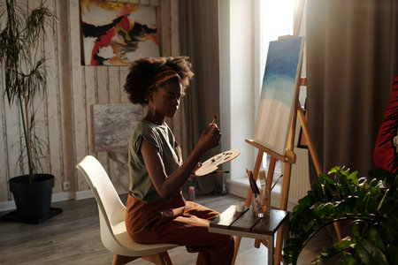 Young artist surrounded by art supplies, creating painting on canvas in sunlit studio with open window and plant decor highlighting her focused expressionの写真素材
