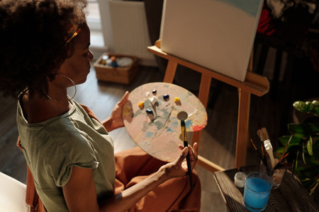 Young African American woman creating art, holding paint palette and brush, working on canvas while seated in well-lit studio during daytime, showcasing artistic processの写真素材