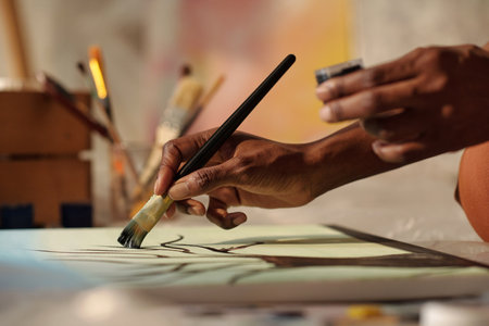 Close-up view of artists hand holding paintbrush while painting on canvas with brushes and tools...