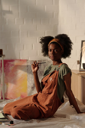 African American woman sitting casually on floor holding paintbrush creating art next to her...
