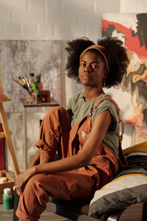 Portrait of young African American woman sitting comfortably in art studio, surrounded by painting...