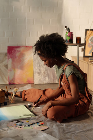 African American woman intensely working on a painting in a bright studio surrounded by art supplies...