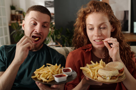 Young modern heterosexual couple eating french fries while sitting next to one another on couch in...