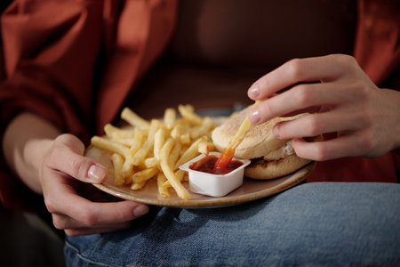 Hands of young unrecognizable woman putting french fries in ketchup in small plastic container while...