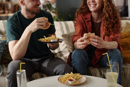 Cropped shot of young man with plate of french fries sitting next to his girlfriend with hamburger while both having junk food for lunchの写真素材