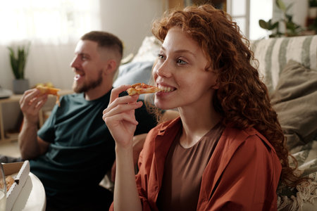 Young smiling woman biting slice of tasty pizza and watching movie on home cinema against her happy boyfriendの写真素材