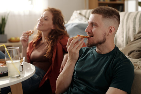 Young man eating takeout pizza and watching movie while sitting in front of camera on the floor against his girlfriendの写真素材