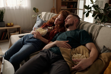 Tired girl and guy with bowls of popcorn in hands sleeping on couch in home environment after watching movie or tv broadcastの写真素材