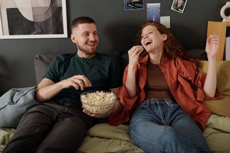Carefree laughing girl eating popcorn while relaxing on bed with her boyfriend with remote control...