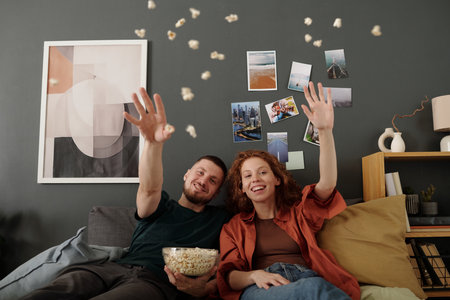 Happy young man and woman throwing popcorn and looking at camera with smiles while sitting on bed and enjoying movieの写真素材