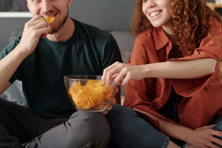 Cropped shot of smiling girl taking crispy potato chips from bowl held by her boyfriend while both sitting in front of cameraの写真素材