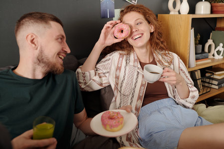 Happy young woman with cup of tea laughing and looking at her boyfriend while holding donut in front of her faceの写真素材