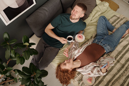 High angle of young laughing couple relaxing on double bed in the morning and having donuts with tea for breakfastの写真素材