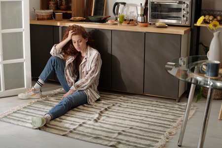 Young serene or upset woman in casual apparel sitting on the floor by kitchen counter with kitchenware and junk foodの写真素材