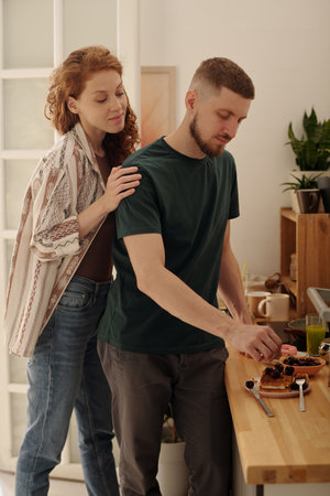 Young affectionate woman embracing her husband or boyfriend cooking dessert with homemade waffles...