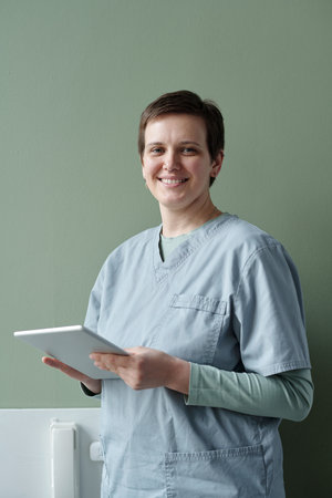 Portrait of smiling nurse holding digital tablet looking at camera in hospital setting wearing...