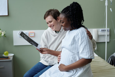 Pregnant woman in hospital gown sharing ultrasound image with another smiling person in hospital...
