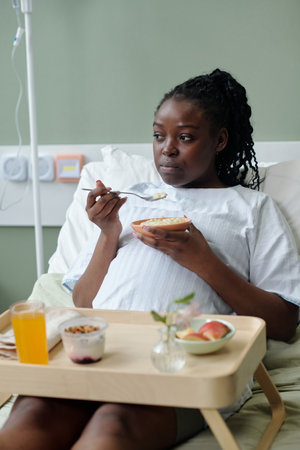 Woman sitting in hospital bed enjoying breakfast while recovering from medical treatment Eating healthy food including yogurt, fruit, and juice with an IV stand next to bedの写真素材