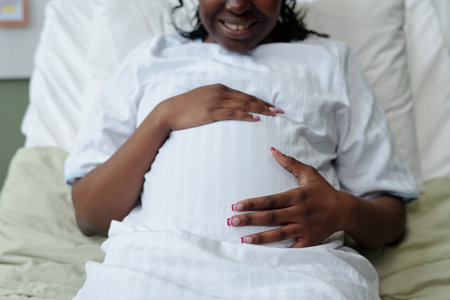Expectant mother smiling while gently touching her pregnant belly, wearing a comfortable white dress and seated indoorsの写真素材