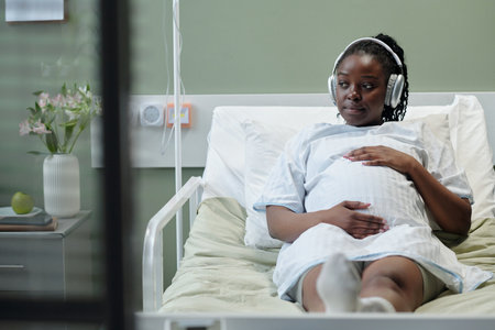 African American woman relaxing in hospital bed with headphones on, enjoying music. Vase with flowers and medical equipment in background creating a calm environmentの写真素材